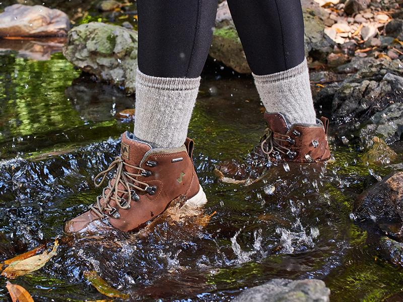 The image features a person walking in water wearing the Thatcher Women's Waterproof Sneakerboots in Toffee.